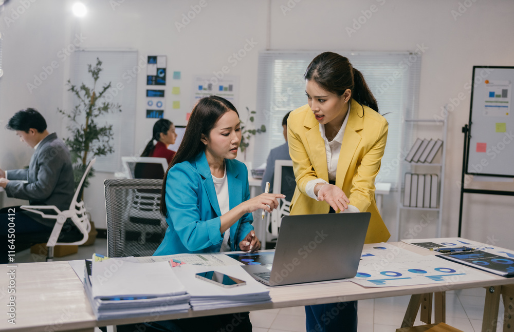 Two asian businesswomen are discussing a project using a laptop in a busy office, analyzing charts and collaborating on a task Two asian businesswomen are discussing a project using a laptop in a busy office, analyzing charts and collaborating on a task