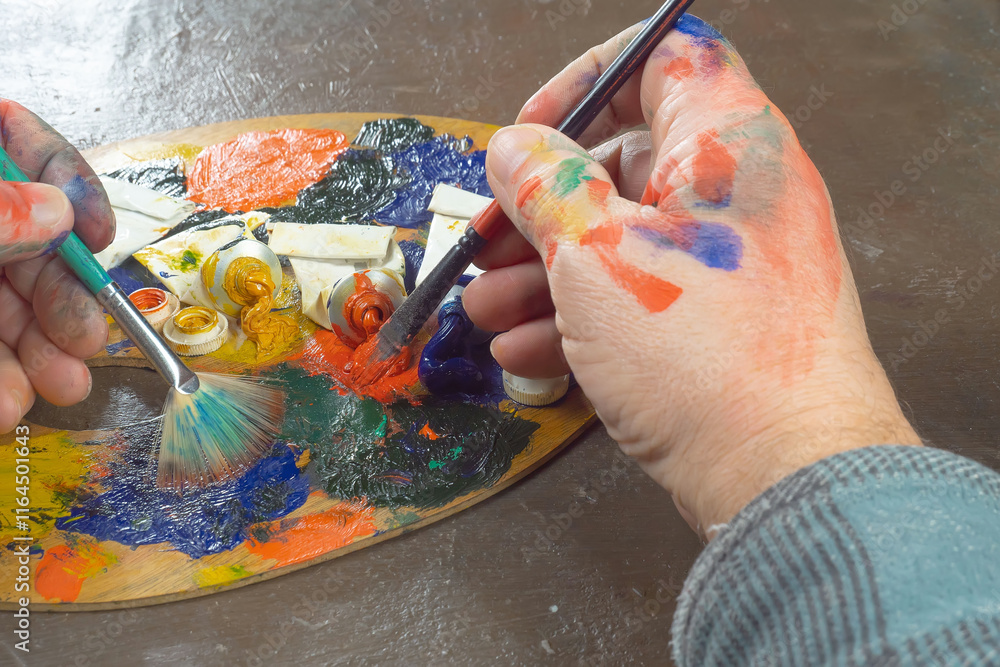 Obraz premium The hands of the artist, work to create creative images in art. Art paints and brushes, applying multi-coloured strokes to the canvas using palette, gouache and watercolour.