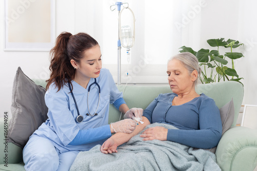 Young friendly nurse or doctor help sick senior elderly woman patient in receiving IV drip infusion and vitamin injection therapy sitting on sofa in medical clinic or at home. Health care concept