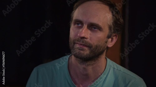 A man gazes upwards with a bored and slightly weary expression, his head resting on his hand. The simple background and indoor setting contribute to the overall mood