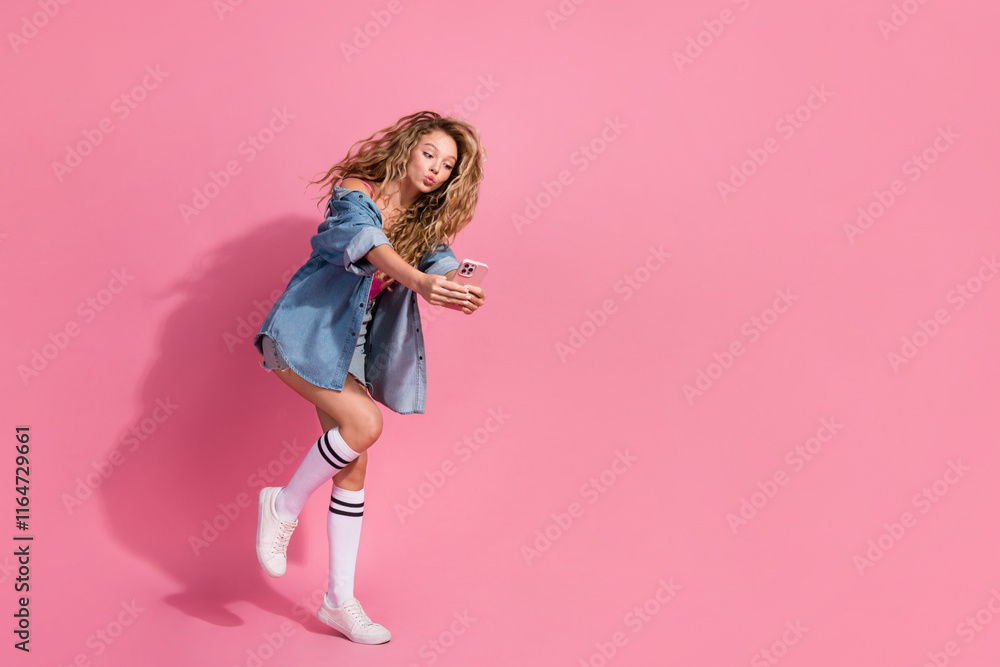 Young woman in stylish denim jacket taking a selfie against a vibrant pink background, showcasing casual and trendy fashion