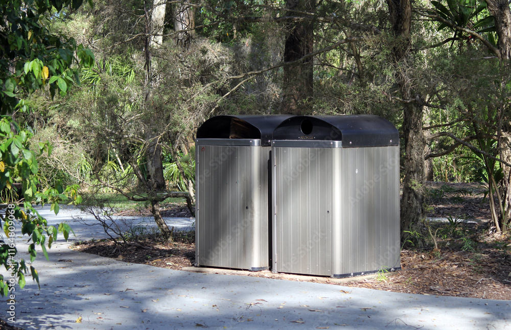 Two metal trash cans bins next to a concrete path in a park with trees Two metal trash cans bins next to a concrete path in a park with trees