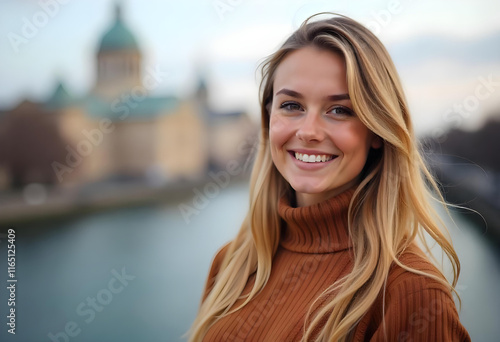 A young Caucasian woman with long blonde hair wearing a brown sweater , smiling and looking at the camera against a blurred cityscape background