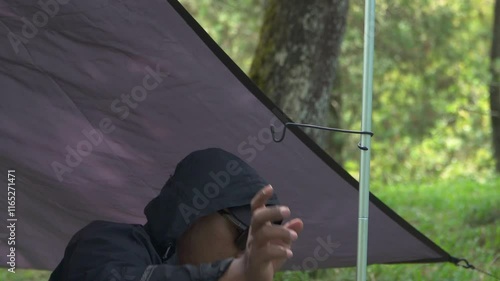 A solo traveler is preparing a flysheet tent in a green and lush pine forest area. Asian man adventuring alone in the jungle and setting up a trap tent from flysheet for bushcraft and camping