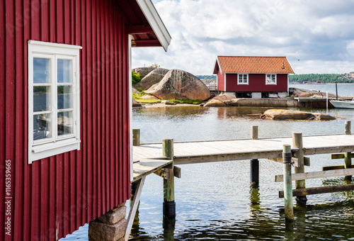 Two red painted boathouses with tiled roofs and a wooden pier on the pink granite coast in the Bohuslän province, Sweden, under a stormy sky on a sunny summer day.