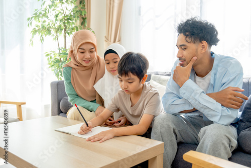 Side and upper view of muslim family with mother, father and daughter sit and cheer little boy for writing in living room of their house. photo