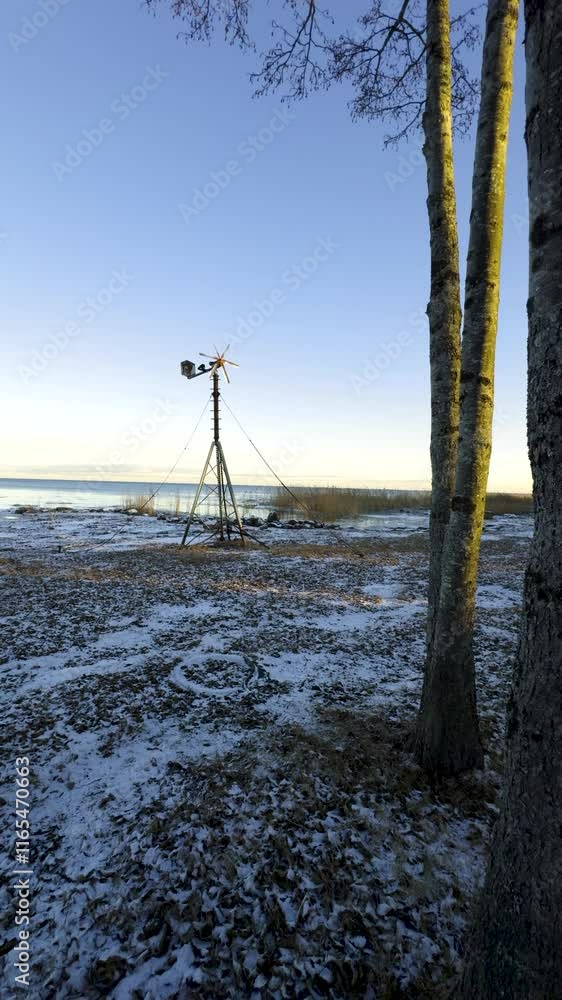 A close-up of a weathered windmill standing on the snowy shore of Lake Ladoga, with calm water and reeds in the background. The sunset light highlights the rustic structure against the winter A close-up of a weathered windmill standing on the snowy shore of Lake Ladoga, with calm water and reeds in the background. The sunset light highlights the rustic structure against the winter