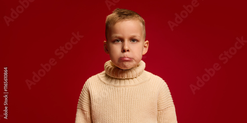 Принти на полотні Boy frowns slightly, creating pouting expression, wearing knitted sweater, standing against deep red background