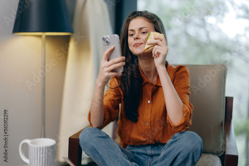 Smiling young woman enjoying a snack while using her smartphone in a cozy indoor setting, embracing relaxation and modern lifestyle with warm tones photo