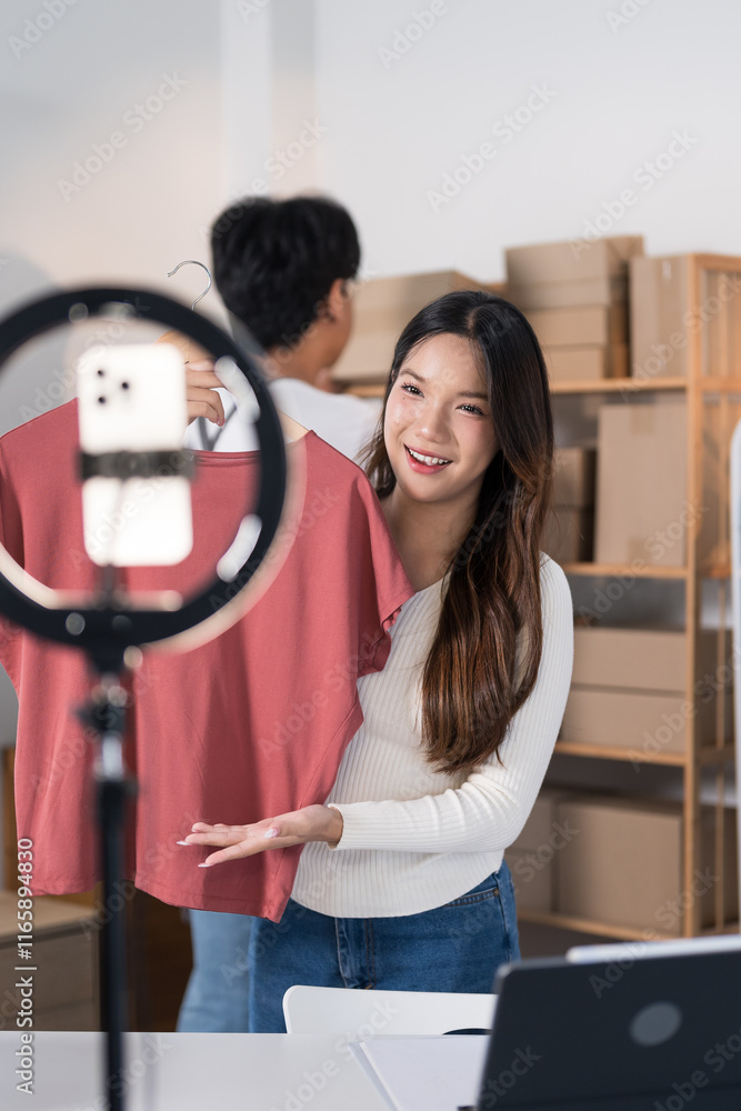 Obraz premium A young woman livestreams showcasing a shirt to her online audience using a smartphone and ring light, with packaging materials and clothing racks in the background