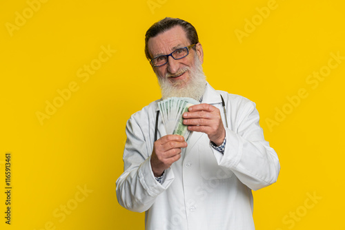 Rich happy mature doctor cardiologist man waving money dollar cash banknotes bills like a fan, success business career, big income wealth. Apothecary pharmacy grandfather isolated on yellow background photo