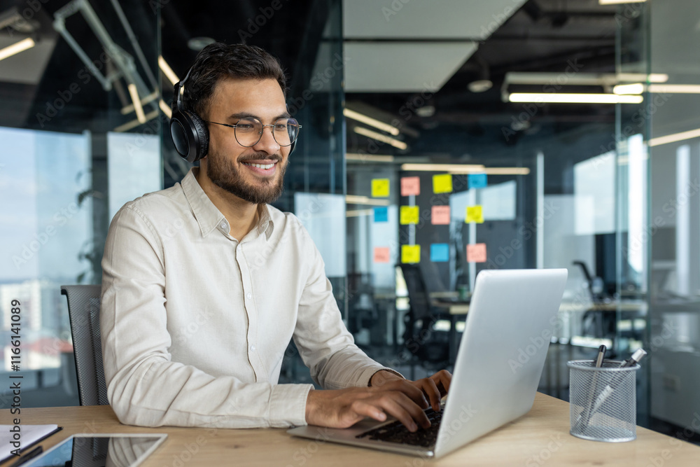 Latin American man using a laptop while wearing headphones in a modern office environment. Captures a professional setting, teamwork planning in the background, and focus on productive work. Latin American man using a laptop while wearing headphones in a modern office environment. Captures a professional setting, teamwork planning in the background, and focus on productive work.