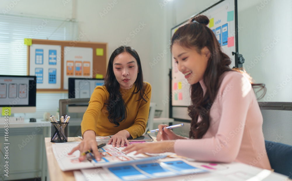 Fototapeta premium Two female ux designers are collaborating on wireframes for a mobile app, reviewing design layouts and user interface elements in their modern office
