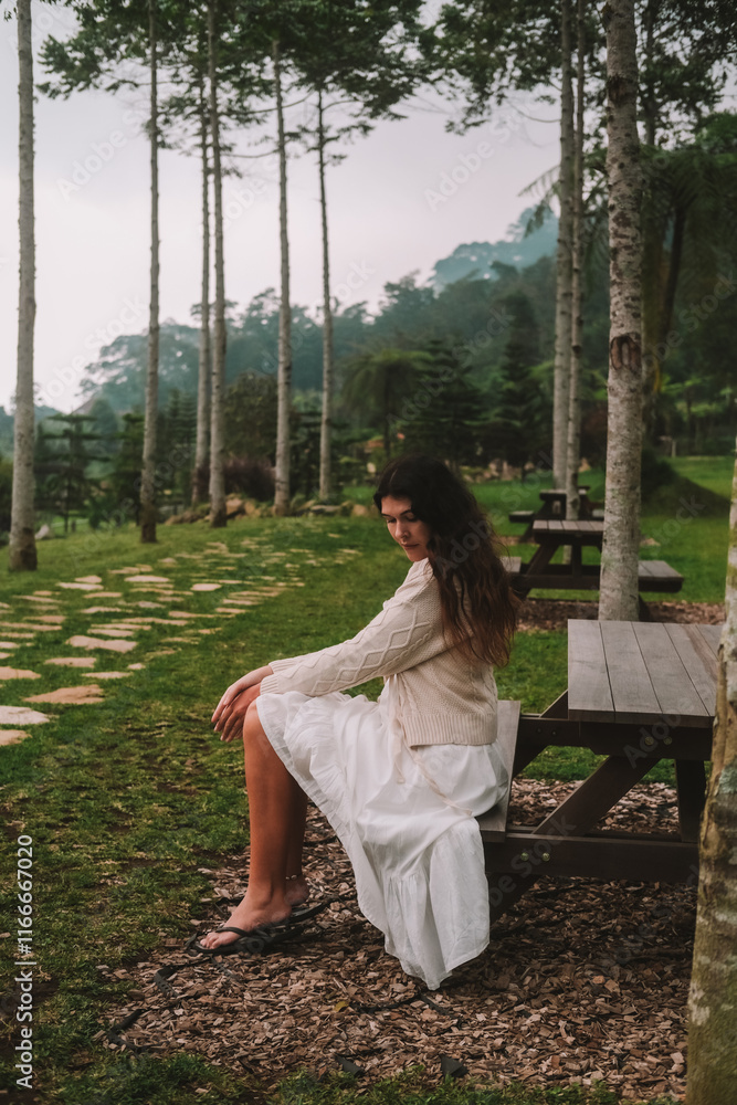 Young woman in white dress sitting on bench on rural farm with green hilly mountains and trees in background. She is relaxing. Ecological outdoor tourism on farm in mountains. Young woman in white dress sitting on bench on rural farm with green hilly mountains and trees in background. She is relaxing. Ecological outdoor tourism on farm in mountains.