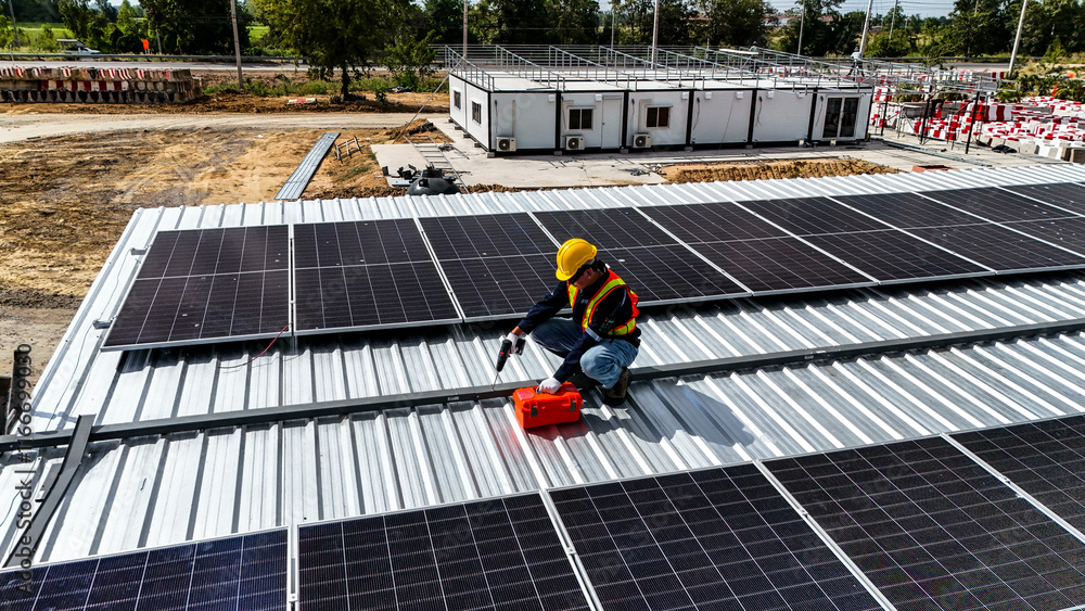 Naklejka premium A worker wearing safety gear installing solar panels on a rooftop with nearby housing units in the background, emphasizing renewable energy, sustainability, and efficient solar power implementation