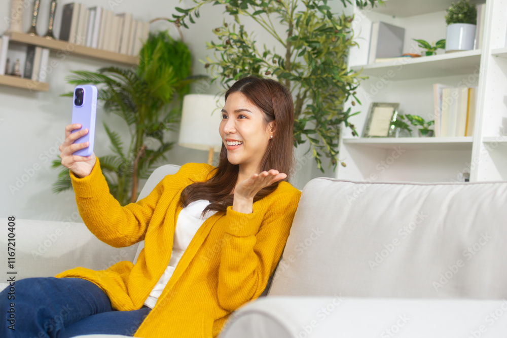 Cheerful young Asian woman making a video call with a friend, sitting on sofa in living room at home. Smiling Asian woman holding smartphone enjoys video call online conversation from apartment room. Cheerful young Asian woman making a video call with a friend, sitting on sofa in living room at home. Smiling Asian woman holding smartphone enjoys video call online conversation from apartment room.