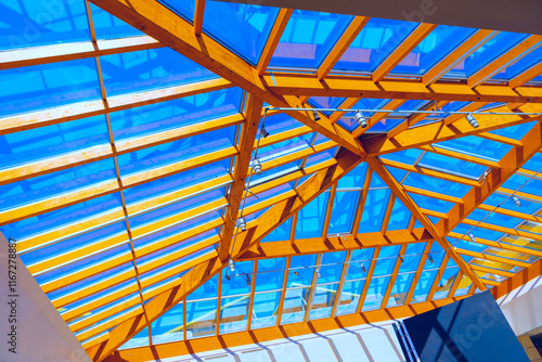 Modern architectural structure with wooden framework supporting glass roof. Vibrant sky visible through glass panels and intricate design of wooden beams make structure architecturally interesting