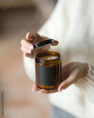 Aroma soy candle in brown glass jar with black lid in woman's hands. Amber minimalist candle packaging design with copy space. Aromatherapy, craft home decor, gift idea, home comfort, relaxation.