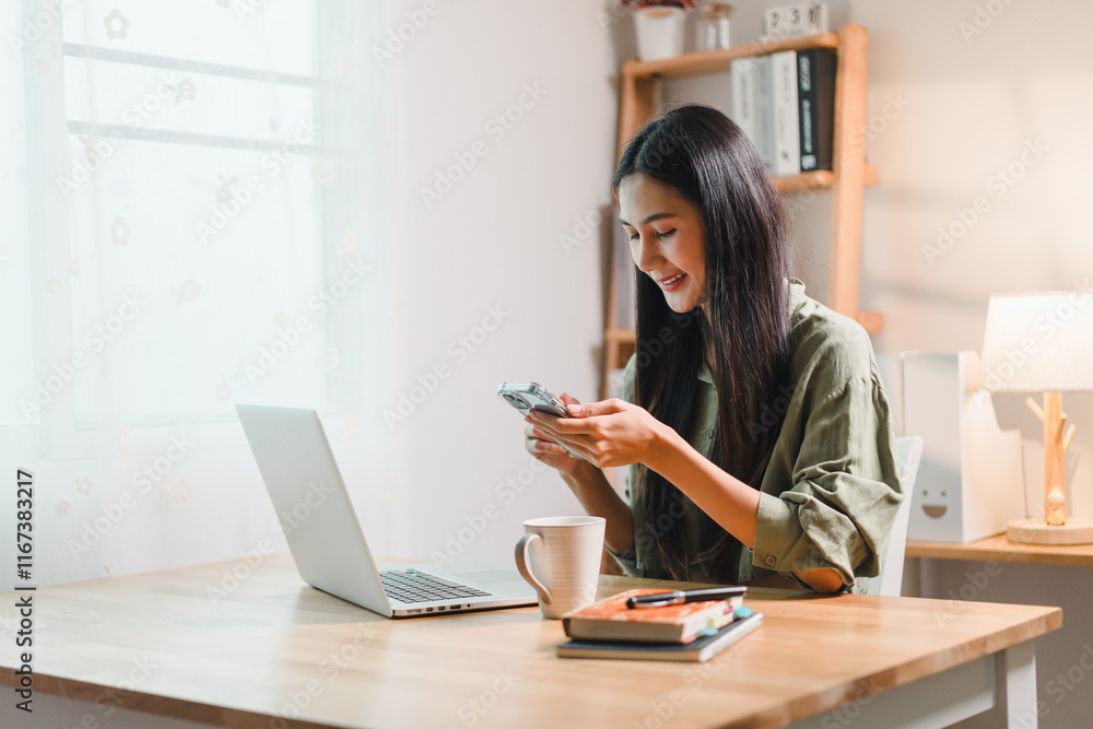 young woman smiling while using her smartphone at wooden desk with laptop, coffee mug, and books in bright home office young woman smiling while using her smartphone at wooden desk with laptop, coffee mug, and books in bright home office