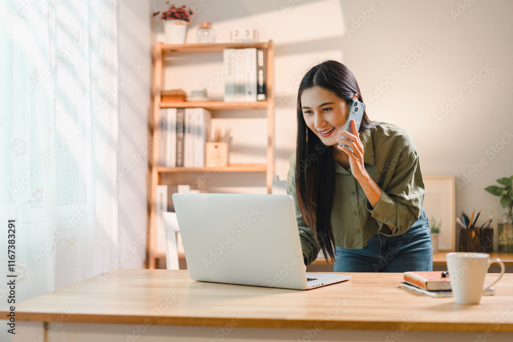 young woman smiling while talking on phone and working on laptop at wooden desk in bright home office with bookshelves and decor in background young woman smiling while talking on phone and working on laptop at wooden desk in bright home office with bookshelves and decor in background