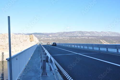 Arched concrete bridge spanning across a tranquil body of water, on the backdrop of green hills and distant mountains. Krk bridge, Croatia