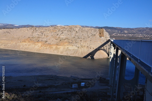 Arched concrete bridge spanning across a tranquil body of water, on the backdrop of green hills and distant mountains. Krk bridge, Croatia
