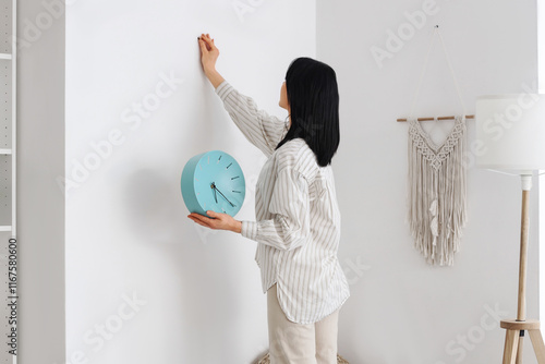 Young woman hanging blue clock on light wall at home photo