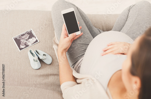 Unrecognizable pregnant woman using smartphone with blank screen, with tiny shoes and sonogram. Young expectant lady surfing net, reading about baby care, buying goods for newborn, copy space