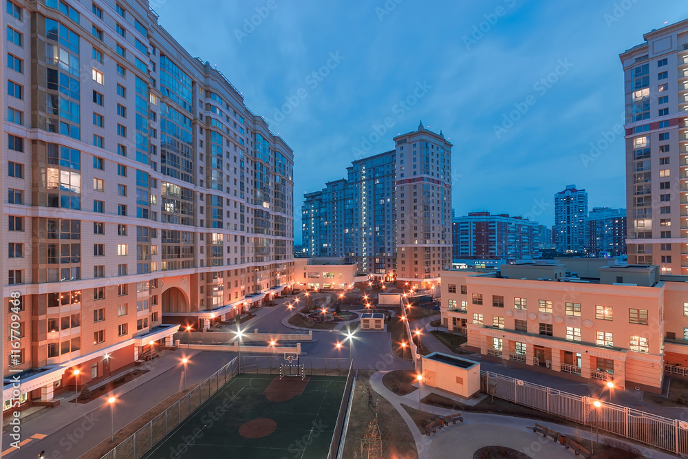 Fototapeta premium The scene captures a modern residential area at twilight, showcasing illuminated apartments and a recreational space with a sports court. The urban landscape is lively and welcoming.