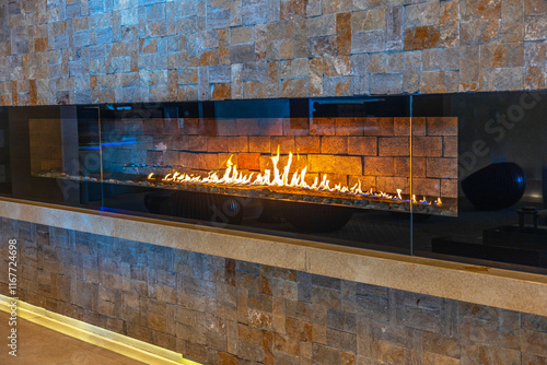 Modern linear gas fireplace with vibrant flames set against textured stone wall in cozy interior space hotel. Mexico.