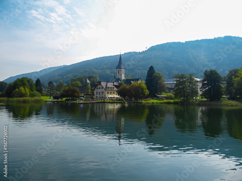 Tranquil lakeside scene with Ossiach Abbey nestled amidst rolling hills of Ossiacher Tauern in Carinhtia, Austria. Panoramic boat tour on Lake Ossiach in summer. Benedictine monastery in Austrian Alps photo