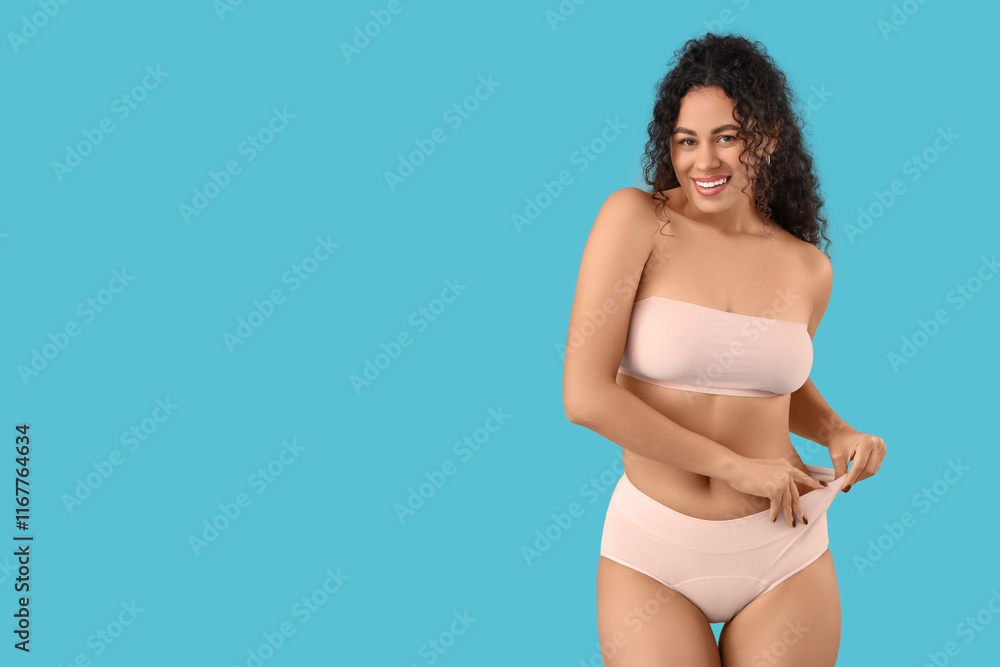 Fototapeta premium Beautiful young happy African-American woman in elegant pink underwear on blue background