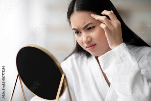 Closeup of upset young asian lady in white bathrobe looking at mirror in bedroom, checking her wrinkles on forehead, copy space. Sad pretty korean woman got irritated with pimples on her face photo