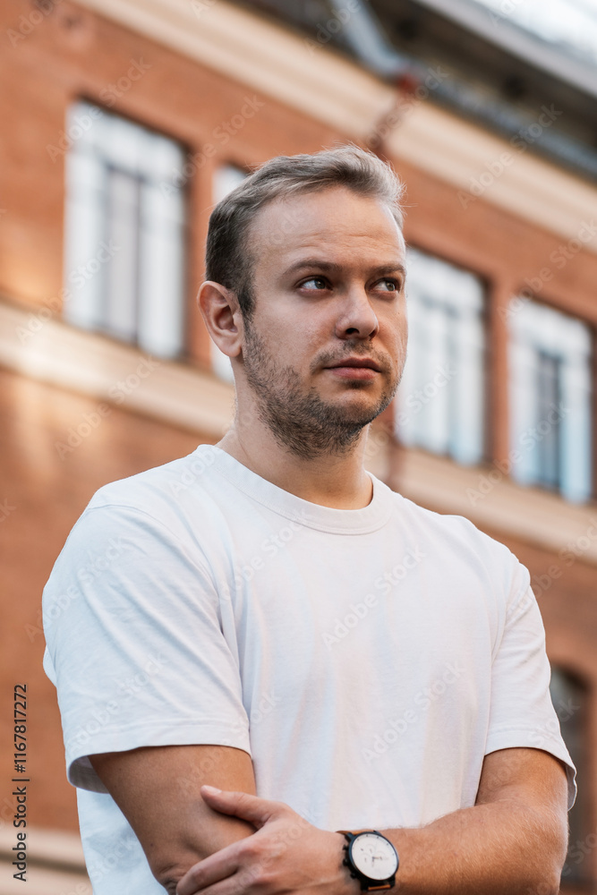 A man with a beard, wearing a white tshirt and a watch, stands in front of a brick building. His chin is raised, revealing a strong jawline A man with a beard, wearing a white tshirt and a watch, stands in front of a brick building. His chin is raised, revealing a strong jawline