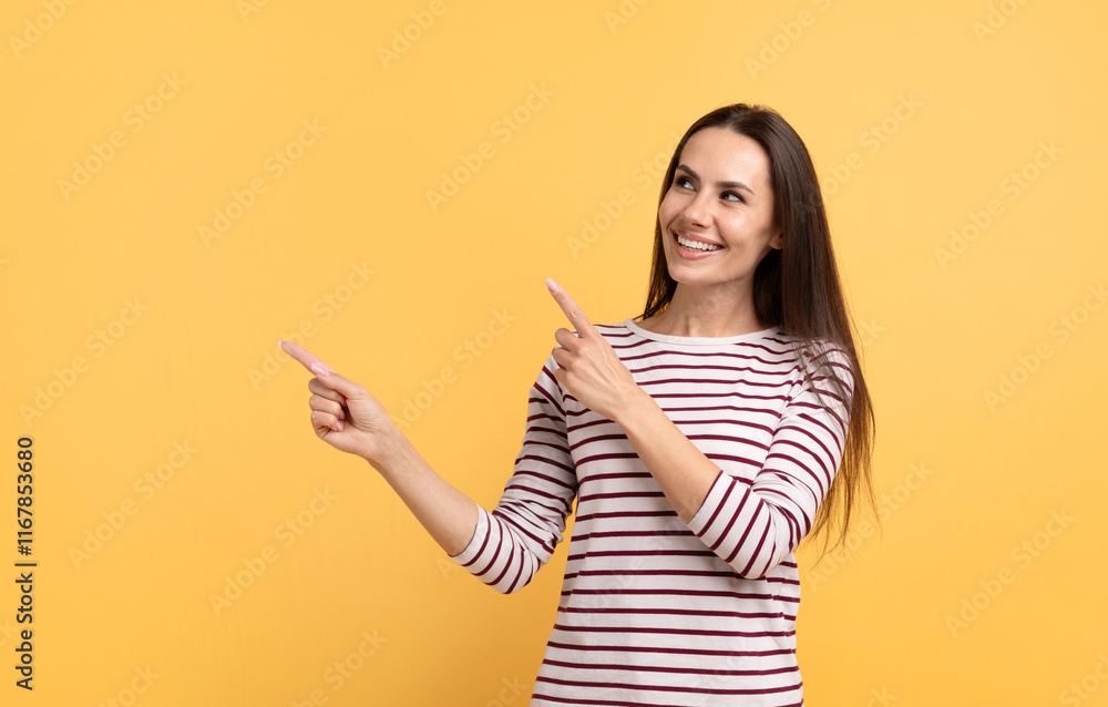 Positive young brunette woman pointing at copy space with both hands and smiling at camera, isolated on yellow studio background Positive young brunette woman pointing at copy space with both hands and smiling at camera, isolated on yellow studio background