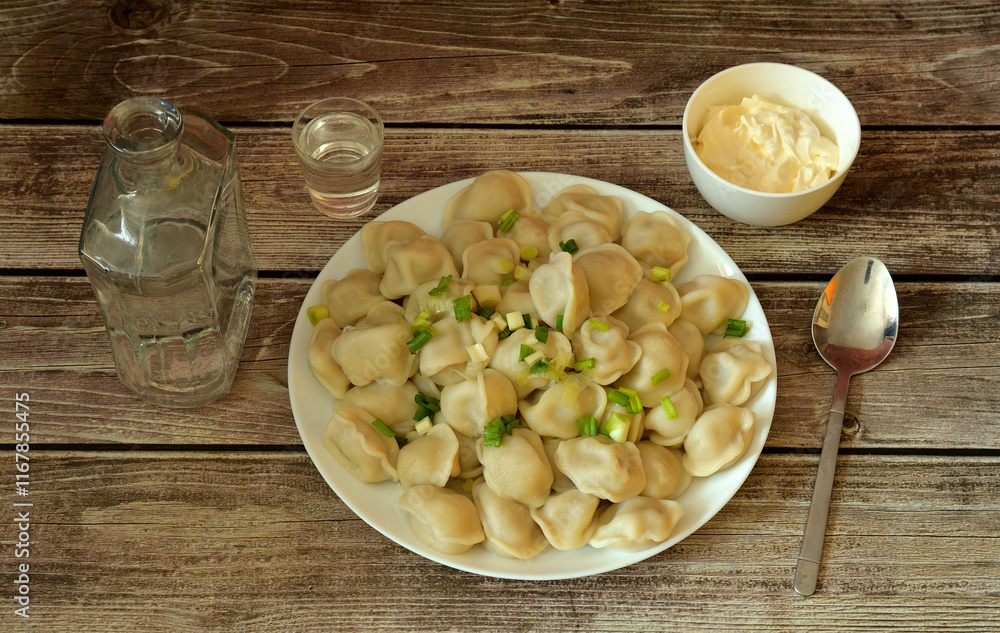 A plate of freshly cooked meat dumplings with sour cream and herbs on a wooden table, a bottle and a shot glass with vodka and a spoon nearby. A plate of freshly cooked meat dumplings with sour cream and herbs on a wooden table, a bottle and a shot glass with vodka and a spoon nearby.