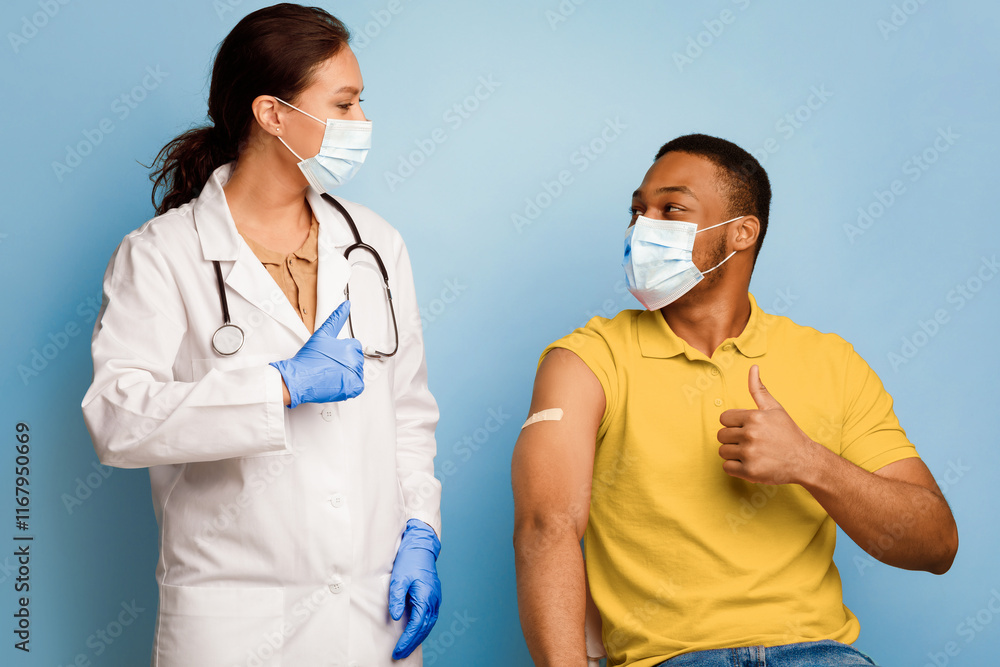 Covid-19 Vaccination. Vaccinated Black Man And Medical Worker Gesturing Thumbs-Up After Successful Coronavirus Immunization Posing Wearing Face Masks In Studio Over Blue Background. Covid-19 Vaccination. Vaccinated Black Man And Medical Worker Gesturing Thumbs-Up After Successful Coronavirus Immunization Posing Wearing Face Masks In Studio Over Blue Background.