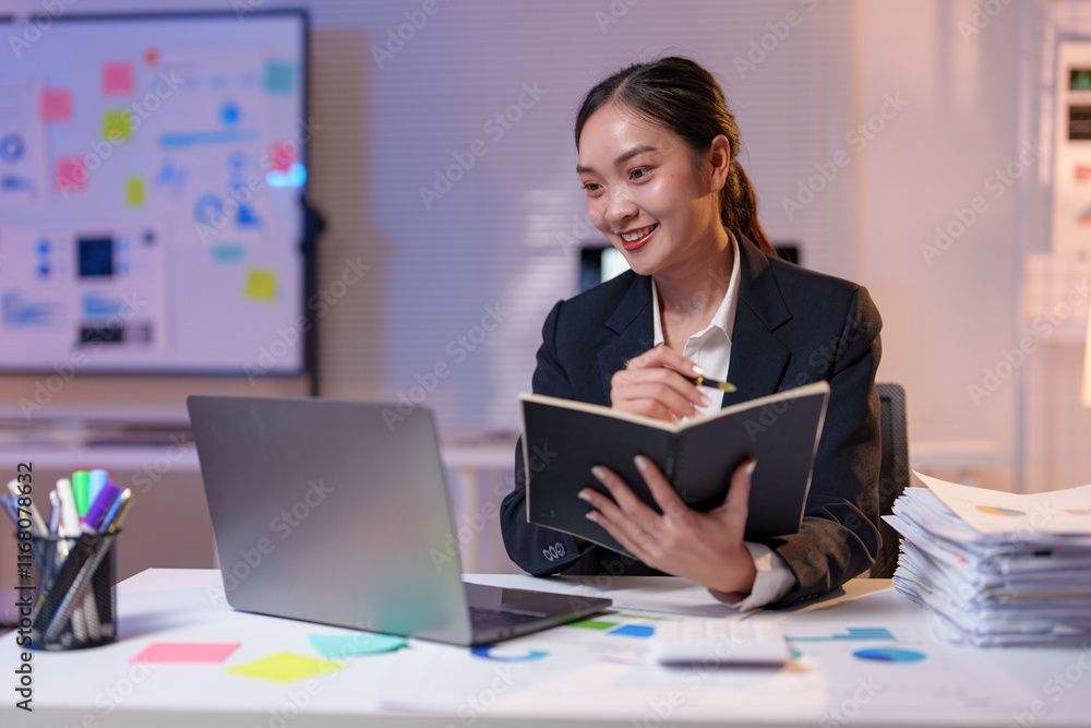 Young professional businesswoman attending a virtual meeting on laptop and taking notes in her personal organizer, working late in a modern office with a whiteboard full of tasks in the background Young professional businesswoman attending a virtual meeting on laptop and taking notes in her personal organizer, working late in a modern office with a whiteboard full of tasks in the background