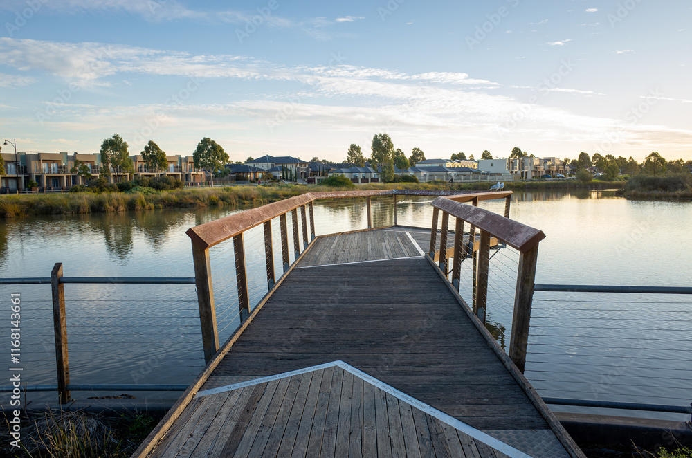 Obraz premium A wooden boardwalk over the still waters of a lake in Tarneit, surrounded by residential houses in the neighborhood. Public recreational amenity in Australia's suburbs. Copy space for design