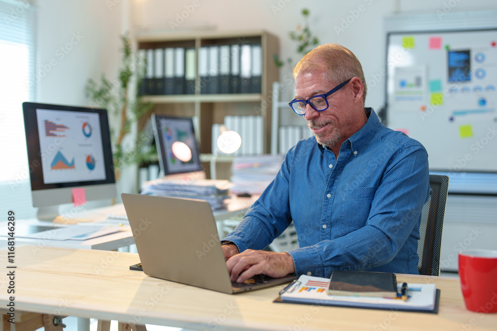 Mature employee concentrating on work while typing on a laptop, surrounded by documents and computer screens displaying charts, exemplifying professionalism in a productive office