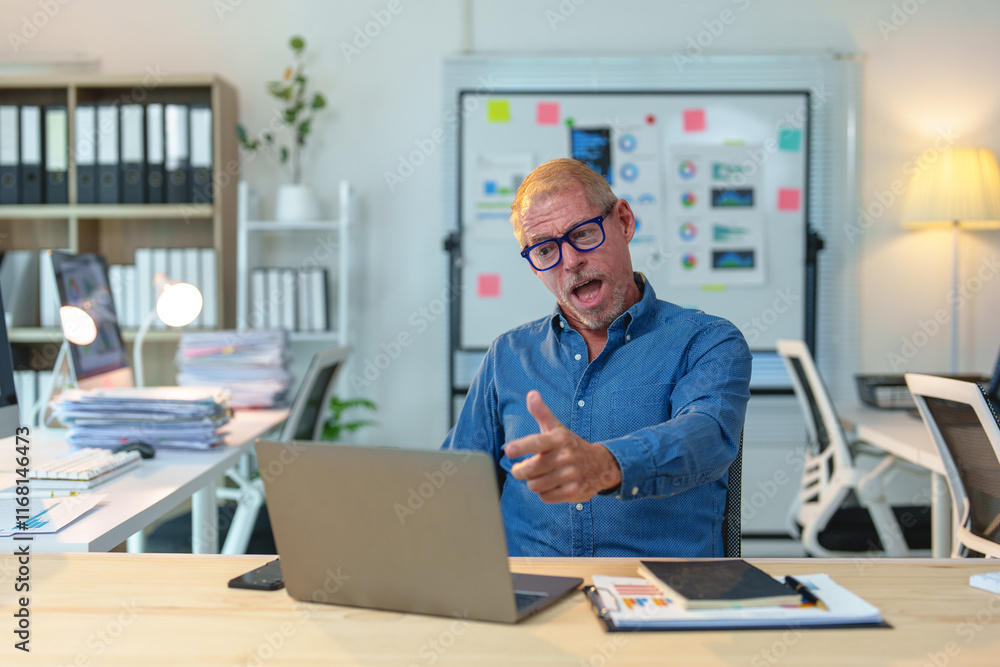 Stressed mature businessman arguing during video conference on laptop in office, pointing his finger at the screen while sitting at his desk with paperwork and documents Stressed mature businessman arguing during video conference on laptop in office, pointing his finger at the screen while sitting at his desk with paperwork and documents
