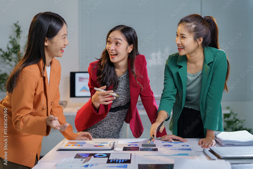 Fototapeta premium Three asian businesswomen discussing marketing strategy in the office, pointing at charts and graphs, sharing insights from reports, smiling and laughing together in a collaborative atmosphere