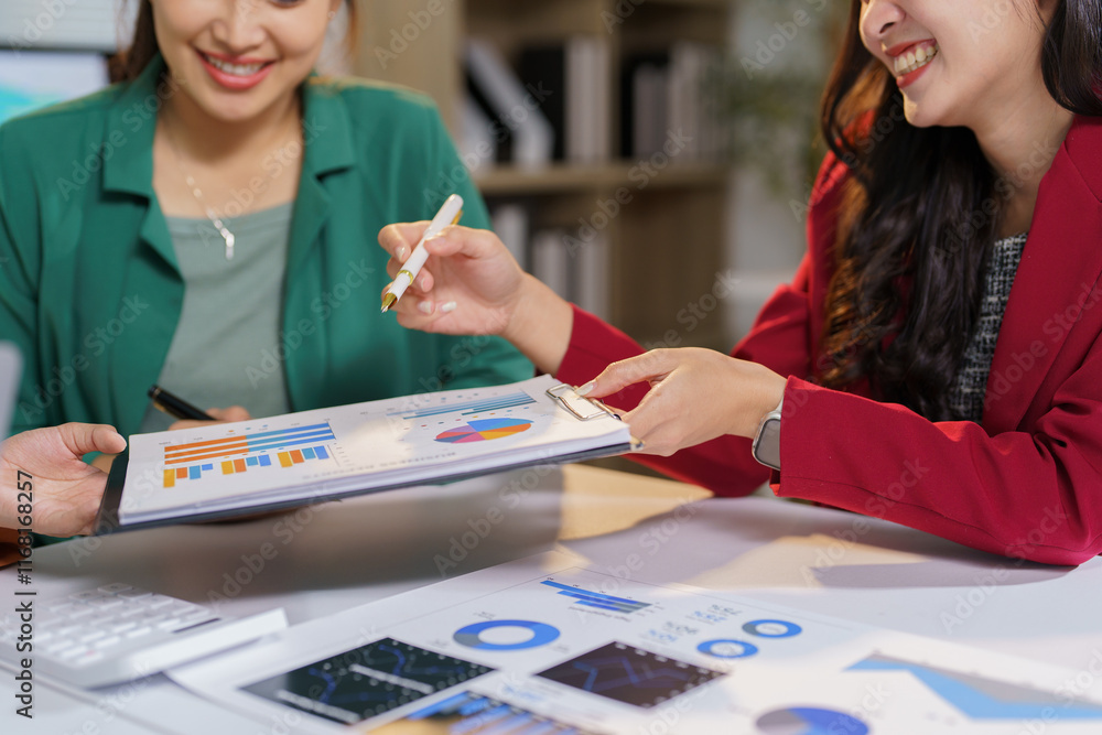 Fototapeta premium Two smiling businesswomen are collaborating on a project, analyzing financial charts and graphs together in a modern office environment, discussing data and strategizing for success