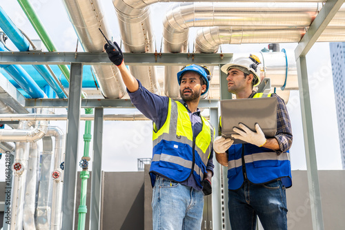 Collaboration, Two young construction workers discuss plans and use laptop computer while reviewing blueprints on a rooftop. They wear safety gear and oversee intricate piping systems in factory photo