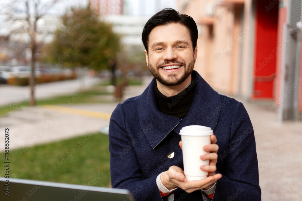 Happy young man in black jacket working with laptop and holding cup with coffee in the city Happy young man in black jacket working with laptop and holding cup with coffee in the city