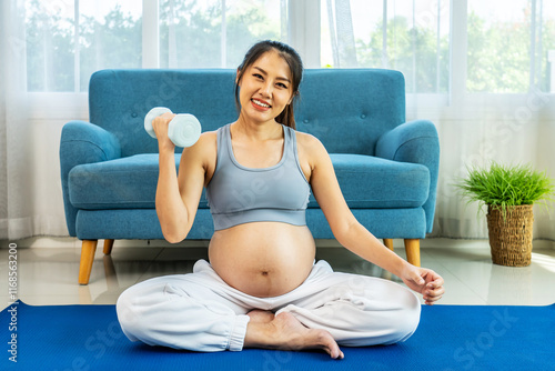 Young pregnant Asian woman about to become a mother, wearing a sports bra, exercising, and sitting lifting dumbbells up and down slowly to flex her arm muscles before giving birth on a yoga mat photo