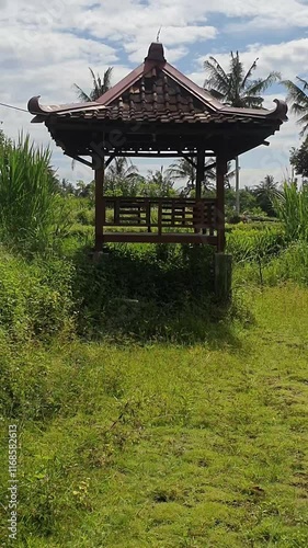 A small wooden pavilion sits in a grassy field. The pavilion is empty and he is abandoned. The surrounding area is lush and green, with tall grass and trees in the background