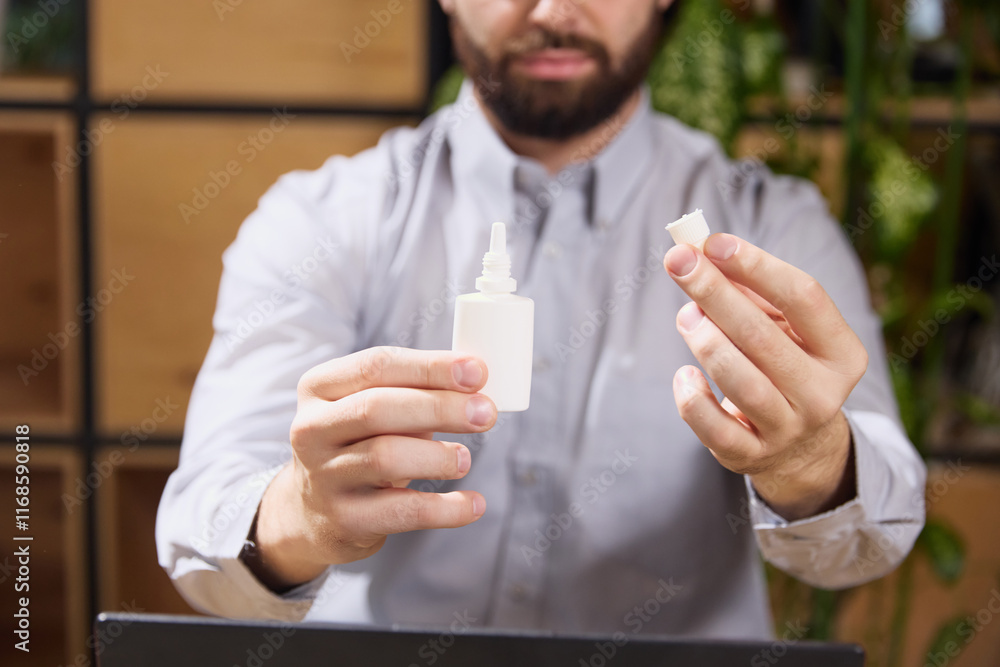 Young man sits at work, and open bottle of nasal spray with negative space to insert your text. Photo with selective focus. Concept of illness, health insurance, pharmaceutical, medicine. Ad. Young man sits at work, and open bottle of nasal spray with negative space to insert your text. Photo with selective focus. Concept of illness, health insurance, pharmaceutical, medicine. Ad.