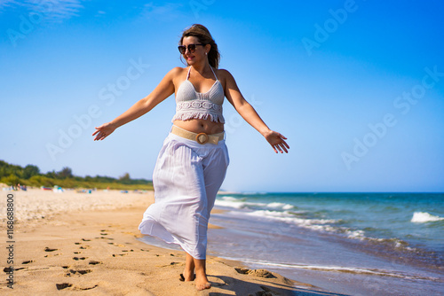 Summertime by sea. Beautiful middle-aged woman walking on sandy beach on sunny summer day. Front view. Beauty and self confidence