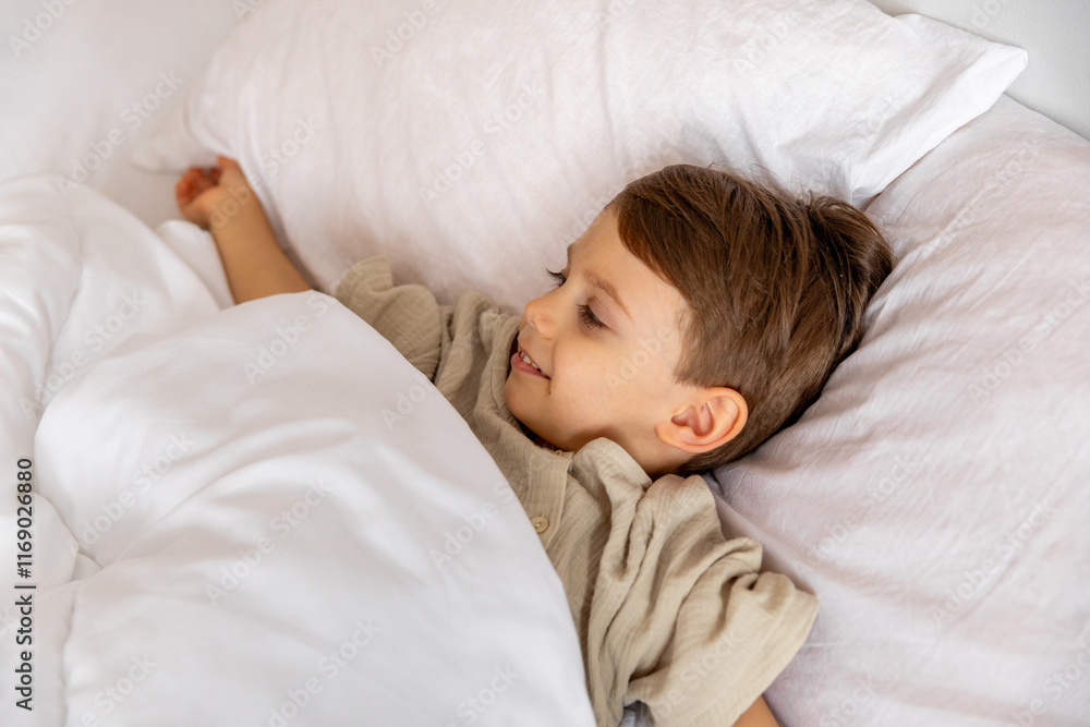 Cheerful little boy lying under white blanket in bed. Child woke up with a good mood in the morning. textiles for the nursery. Have a nice day. Motherhood concept. High quality photo. Copy space Cheerful little boy lying under white blanket in bed. Child woke up with a good mood in the morning. textiles for the nursery. Have a nice day. Motherhood concept. High quality photo. Copy space